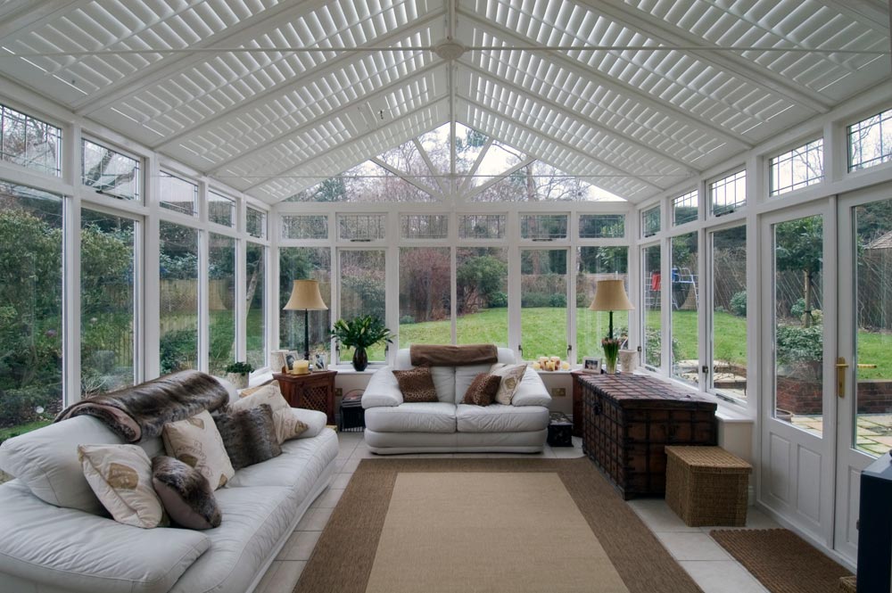 White plantation shutters lining the roof of a patio area surrounded with windows, white leather couches, a beige rug and dark wood patio furniture.