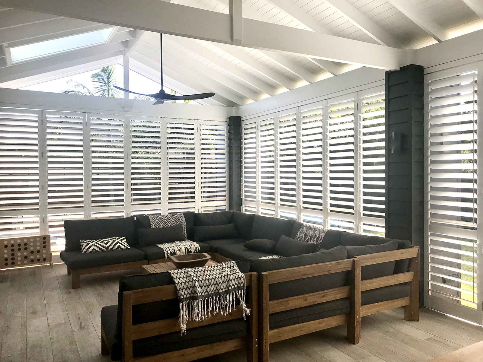 White plantation shutters lining the roof of a patio area surrounded with windows, white leather couches, a beige rug and dark wood patio furniture.